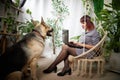 happy beautiful girl in chair using her laptop with cute big dog. Adult middle aged woman with shepherd. Friendship and Royalty Free Stock Photo