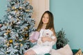 Happy beautiful girl holding a gift box and a stuffed bear toy and sitting near the Christmas tree on the sofa Royalty Free Stock Photo