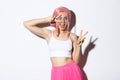 Happy beautiful girl in pink wig, dressed-up for halloween party, showing peace signs, smiling and winking coquettish at Royalty Free Stock Photo