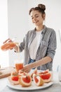 Happy beautiful girl pouring grapefruit detox smoothie in glass smiling over white wall. Healthy diet nutrition.