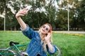 Happy beautiful girl in stylish clothes and sunglasses sits on the grass in the park and takes selfie on the background bike.Young Royalty Free Stock Photo