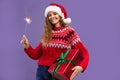 Happy beauty girl wears Santa hat holds gift box and sparkler on purple background. Christmas party Royalty Free Stock Photo