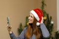 Happy beautiful woman against Christmas tree at home.Cheerful positive lady in santa claus hat using smartphone app call virtual Royalty Free Stock Photo