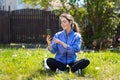 A happy beautiful woman is sitting cross-legged on the grass, using a mobile phone. In the background is a fence. Sunny day, Royalty Free Stock Photo