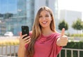 Happy beautiful woman thumbs up with smartphone in her hand and modern city on background. Positive businesswoman smiles at camera Royalty Free Stock Photo