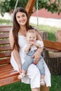 Happy beautiful woman, young mother playing with her adorable baby son and sitting on wooden swing, cute little boy, enjoying Royalty Free Stock Photo