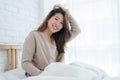 Happy beautiful young Asian woman waking up in morning, sitting on bed, stretching in cozy bedroom, looking through window. Royalty Free Stock Photo