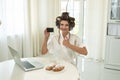 Happy beautiful young brunette in hair curlers holding bank card having successful online shopping on her laptop while drinking
