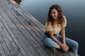 happy beautiful young girl with curly hairs in fashionable blue jeans and yellow blouse sits on a wooden pier Royalty Free Stock Photo