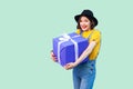 Happy beautiful young girl in fashionable hipster wear in denim overalls and black hat standing and holding big heavy gift box Royalty Free Stock Photo