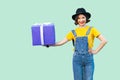 Happy beautiful young girl in hipster wear in denim overalls and black hat standing and holding big heavy gift box with toothy
