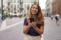 Happy beautiful young student girl looking at camera and holding mobile phone outdoor with blurred city background. City lifestyle Royalty Free Stock Photo