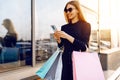 Happy beautiful young woman in sunglasses, with shopping bags, uses a mobile phone, standing in the city against the background of Royalty Free Stock Photo