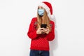 Happy young woman, wearing red santa claus hat and medical protective mask on face, with mobile phone, isolated on white Royalty Free Stock Photo