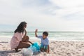 Happy biracial mother and son giving high-five while collecting garbage in bag at beach against sky Royalty Free Stock Photo