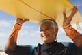 Happy biracial senior woman carrying yellow surfboard over head at beach during sunny day