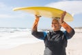 Happy biracial senior woman enjoying retirement while carrying surfboard on head at sunny beach Royalty Free Stock Photo