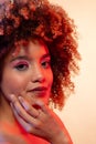 Happy biracial woman with curly hair in pink eyeshadow and lipstick touching face in red light Royalty Free Stock Photo