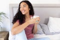 Happy biracial woman holding cup of coffee sitting on bed at sunny home Royalty Free Stock Photo