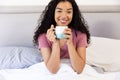 Happy biracial woman holding cup of coffee sitting in bed at sunny home Royalty Free Stock Photo