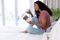 Happy biracial woman reading book and holding cup of coffee sitting on bed at sunny home Royalty Free Stock Photo