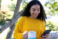 Happy biracial woman in yellow sweater holding cup of coffee and using smartphone on sunny terrace Royalty Free Stock Photo