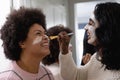 Happy biracial young woman with beauty cream on face applying face pack to female friend at home Royalty Free Stock Photo