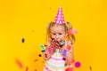 Happy birthday child girl in pink cup blowing confetti on colored yellow background. Celebration, childhood. Royalty Free Stock Photo