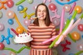 Happy birthday. Festive female. Happy woman with brown hair wearing striped dress holding cake, blowing party horn, celebrating