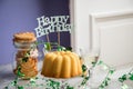 Happy birthday ring cake, cookies, biscuits, muffins and champagne with clover decoration on marble table and lilac background