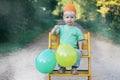 Happy birthday. Smiling little child boy in paper crown with balloons sitting on chair outdoor. Cute toddler celebrating birthday