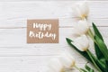 happy birthday text sign on stylish craft greeting card and tulips on white wooden rustic background. flat lay with flowers and g