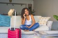 Happy black afro American woman with suitcase talking on mobile phone sitting on sofa couch leaving for holidays trip in tourism a Royalty Free Stock Photo