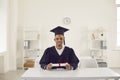 Happy black boy student university graduate in mantle and bonet sitting at desk with diploma Royalty Free Stock Photo