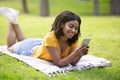 Happy black girl lying on picnic blanket with cellphone at park, communicating with friends or browsing internet