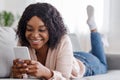 Happy black girl surfing internet on smartphone while lying on couch at home