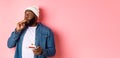 Happy Black hipster celebrating birthday, blowing party whistle, holding bday cake with candle, standing over pink