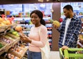 Happy black lady checking shopping list on mobile phone while choosing fruits with her boyfriend at supermarket Royalty Free Stock Photo