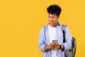 Happy black male student texting on phone with backpack on yellow