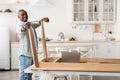 Happy black man measuring the width between legs of wooden table, watching online instructions on laptop in kitchen