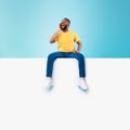 Happy black man sitting on white blank placard, shouting, announcing huge sale on blue background, mockup