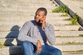 Happy black man talking on smart phone sitting on stairs. Young man in jeans and grey sweater. Concept of communication Royalty Free Stock Photo