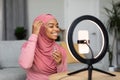 Happy black muslim female beauty blogger making video on smartphone set on ring lamp, showing new facial serum Royalty Free Stock Photo