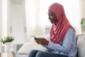 Happy black muslim woman surfing internet on smartphone while resting at home Royalty Free Stock Photo