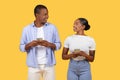 Happy Black spouses with cellphones, sharing smiles over a yellow background