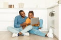 Happy black spouses using laptop and looking for recipes on Internet, sitting on floor in kitchen interior, copy space Royalty Free Stock Photo