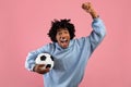 Happy black teenage guy with soccer ball rooting for his favorite football team on pink studio background Royalty Free Stock Photo