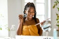 Happy black woman applying makeup at home, lady sitting in front of mirror, holding eyeshadow palette and brush Royalty Free Stock Photo