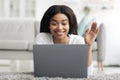 Happy black woman making video call on laptop at home, waving and smiling to screen while lying on floor in living room Royalty Free Stock Photo