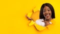 Happy black woman pointing at copy space, presenting huge sale or promo, looking out of hole in torn orange paper